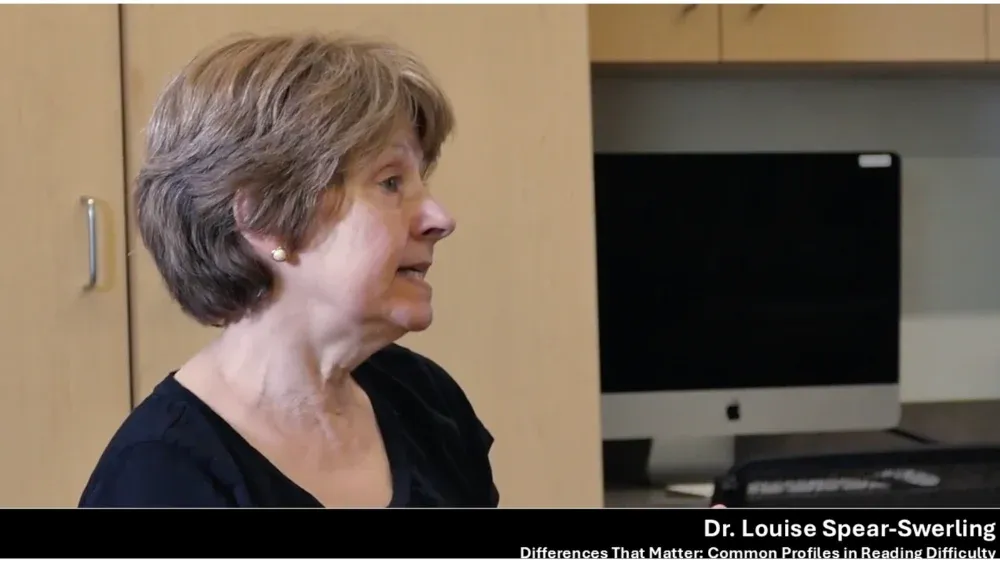 Elderly woman with short brown hair, wearing a black top and pearl earrings, looking to the left.