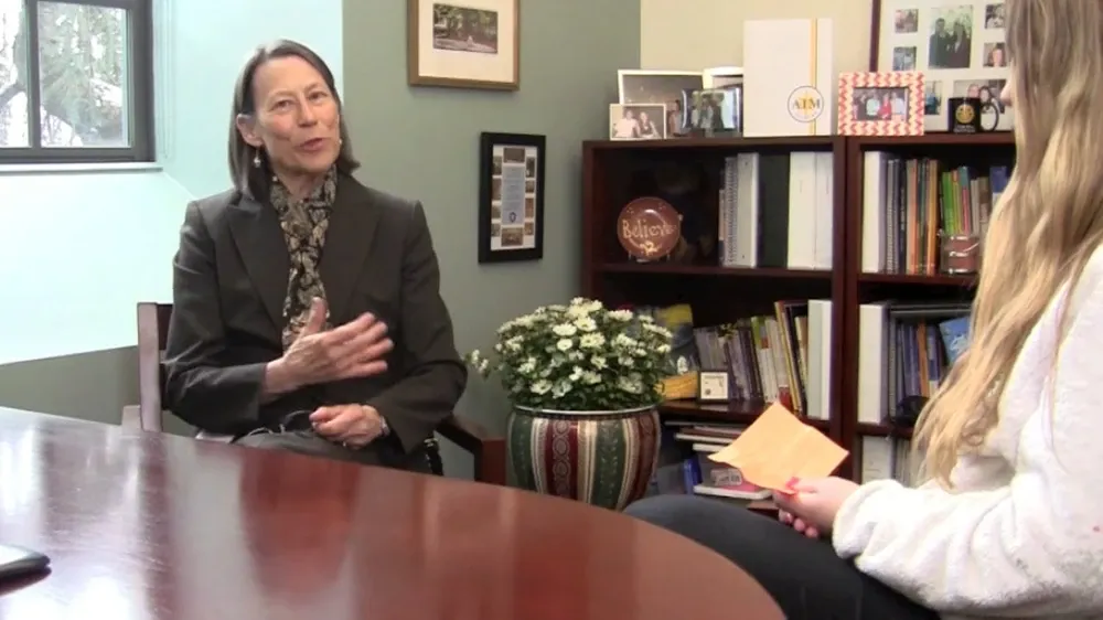 A woman in a dark suit speaks animatedly, gesturing with her left hand while facing a younger woman holding a card.