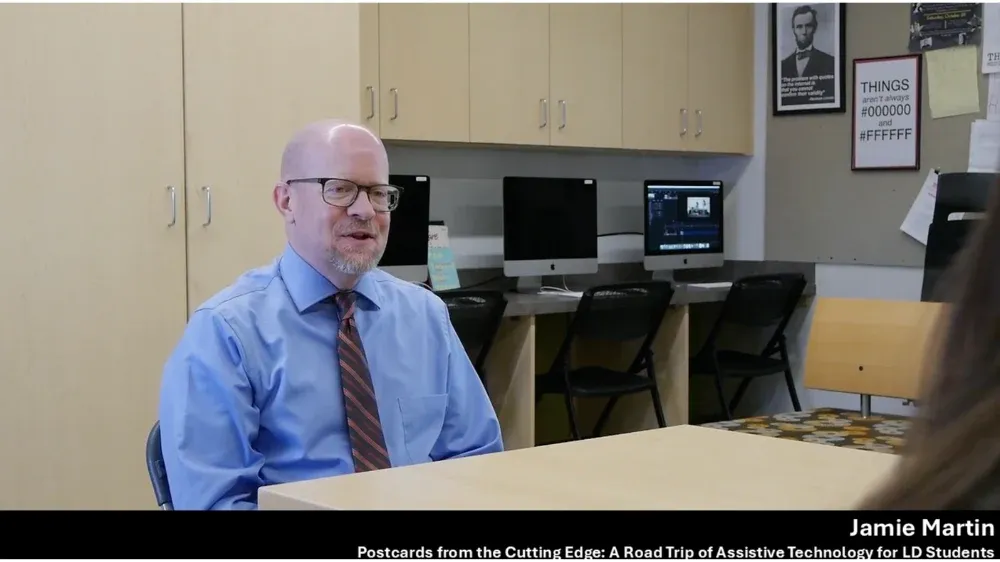 Bald man with glasses and a blue shirt with a striped tie speaks at a table.