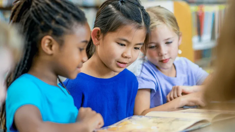 Three diverse young girls collaboratively read an open book in a library setting.