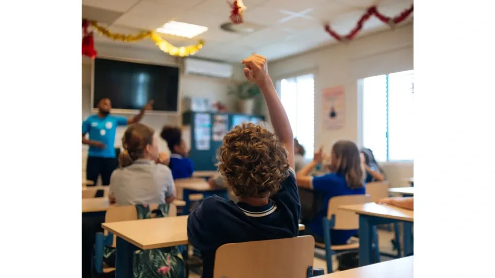 A student with curly hair raises their hand in a classroom while a teacher gestures.