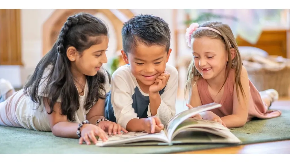 Three diverse children lie on the floor, looking at an open book together.